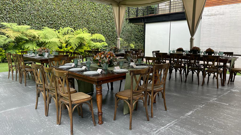 Montaje de evento al aire libre con sillas de madera doble cruz y mesas de madera con patas redondas, decoradas con arreglos florales y vajilla elegante y copas azules aqua. Ideal para eventos y renta de mobiliario.