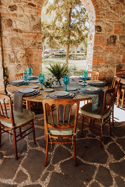 Montaje de mesa al aire libre con sillas de madera Versalles y mesa redonda de madera, decorada con copas azules y vajilla blanca. Ideal para renta y venta en eventos especiales.