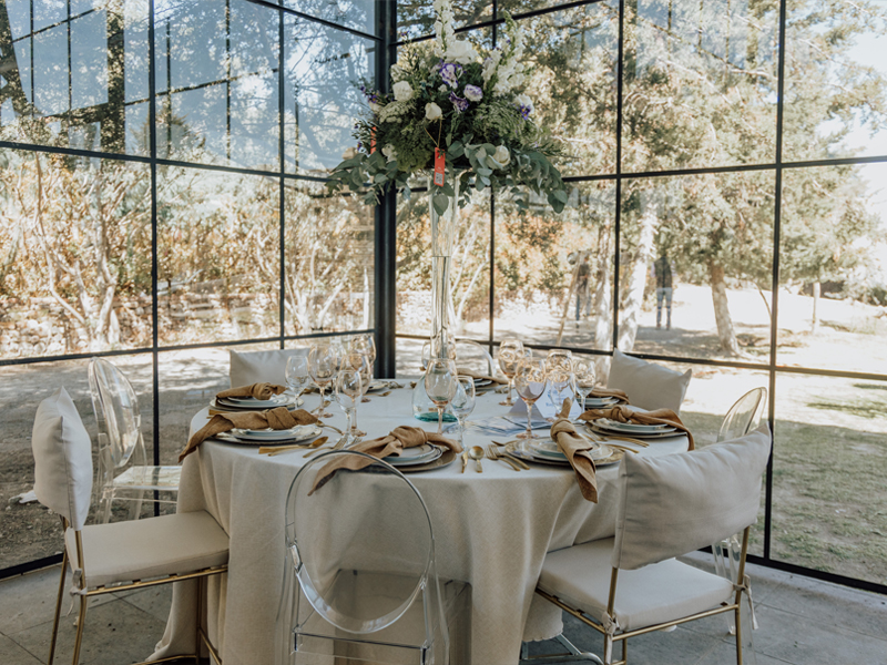 Montaje de una mesa elegante para un evento, con un mantel beige, sillas transparentes y arreglos florales altos en un entorno acristalado con vistas a la naturaleza.