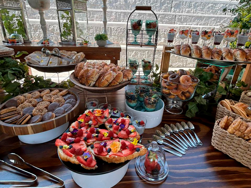 Mesa de catering con una variedad de postres y bocadillos, incluyendo croissants, galletas, frutas frescas con flores comestibles, y vasos de yogur con fruta. La mesa está decorada con elementos rústicos y naturales, creando una presentación elegante y apetecible.
