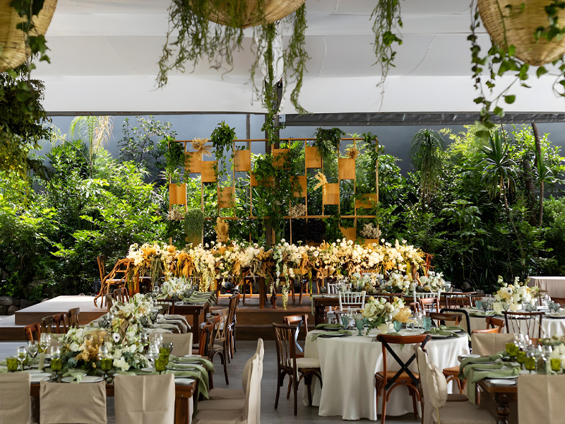 Montaje de evento al aire libre con decoración abundante de flores blancas y amarillas. Mesas redondas y rectangulares están dispuestas con manteles claros, copas verdes y servilletas verdes. El fondo está decorado con vegetación y una estructura de madera con plantas colgantes, ideal para un ambiente natural y elegante.