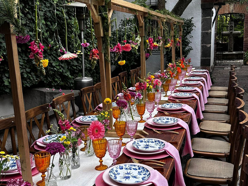 Montaje colorido y festivo para un banquete al aire libre, con una mesa larga decorada con platos de diseño floral azul, copas de colores y arreglos florales colgantes bajo una pérgola de madera.